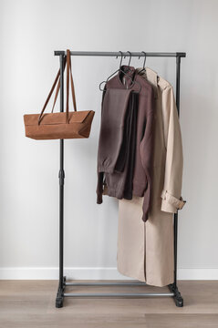 Minimal aesthetic fall fashion capsule wardrobe. Clothing rack with brown clothes, beige trench coat and caramel brown sueded bag, neutral beige empty room interior background
