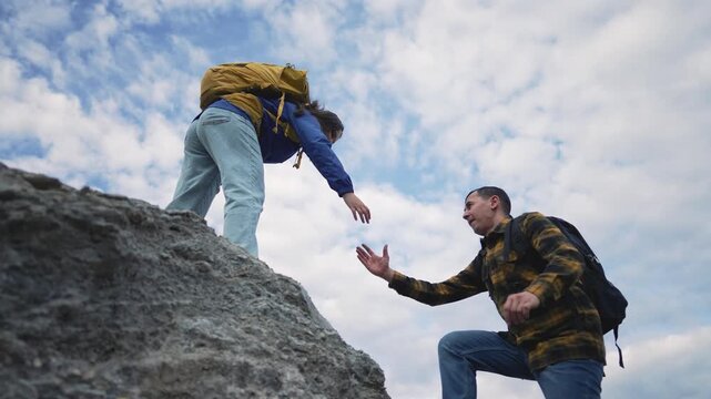 Hiker helping partner climb steep rocky cliff outdoors during mountain adventure. Man assists woman climbing up rock formation. Teamwork and support between hikers on challenging climb.