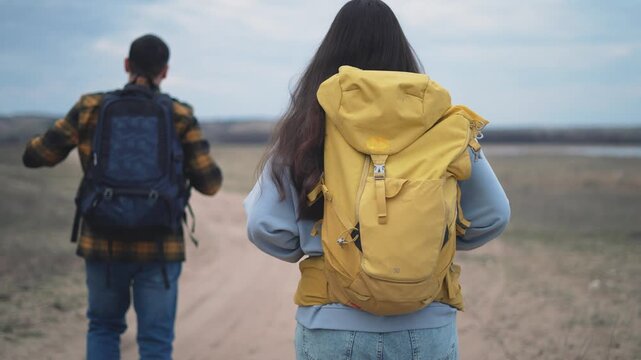 Two young backpackers with colorful backpacks walking along desert trail exploring vast wilderness landscape. Adventure travelers hiking on sandy path through remote arid terrain at sunset.