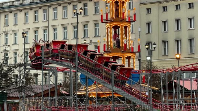 A red sleigh roller coaster moving through a brightly lit Christmas market in a city square, capturing the joyful atmosphere of holiday festivities and winter amusement traditions