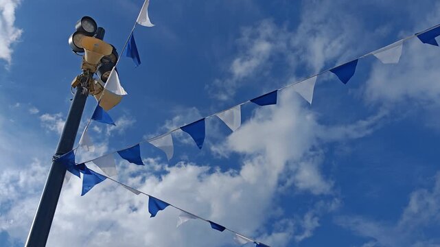Blue and white triangular flags hang from a street lamp against a cloudy sky, festive outdoor decoration, public celebration, summer event, community spirit.