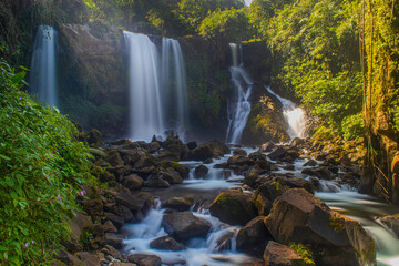 Long exposure shot of beautiful multi-tier waterfalls cascading into a rocky river bed surrounded by lush green tropical forest and warm morning sunlight. © Yudha