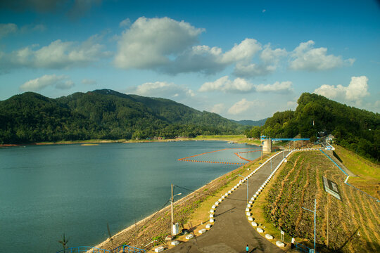 Scenic wide angle view of Sempor Reservoir surrounded by green hills and mountains with sunlight reflecting on the water under a cloudy sky in Kebumen, Central Java, Indonesia.