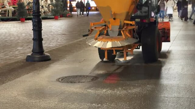 A mechanical salt spreader vehicle driving on a paved urban plaza to deice the ground, maintaining safety during winter season by preventing slippery surfaces for pedestrians in city