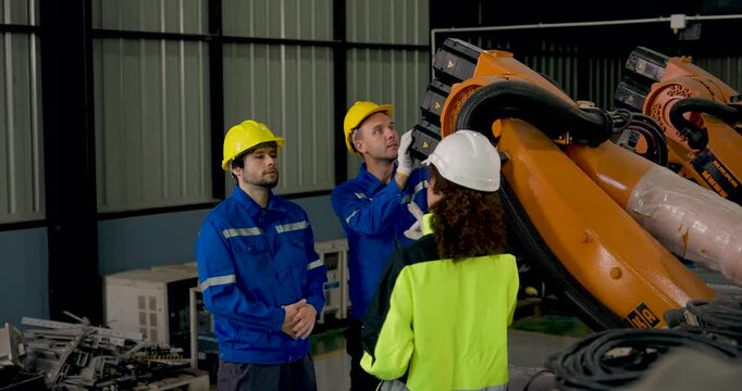 Adult male and female technicians gesture toward industrial robotic arm, examining upper mechanical sections and discussing maintenance procedure during collaborative inspection in factory workshop.