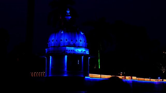 Turquoise and gold illumination brightens the nighttime garden. Swaying palms line the lit promenade. Serene views grace Fateh Sagar Lake in Udaipur, Rajasthan.