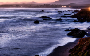 Fototapeta premium Long exposure captures the movement of ocean waves along a rocky coastline during twilight, with distant lights visible on the misty hills. Big Sur, United States.
