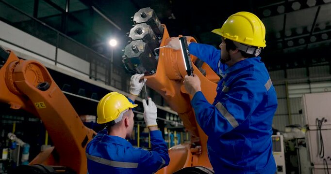 Two adult male technicians inspect industrial robotic arm, moving inspection light across mechanical joint while checking equipment condition during collaborative maintenance in factory workshop.