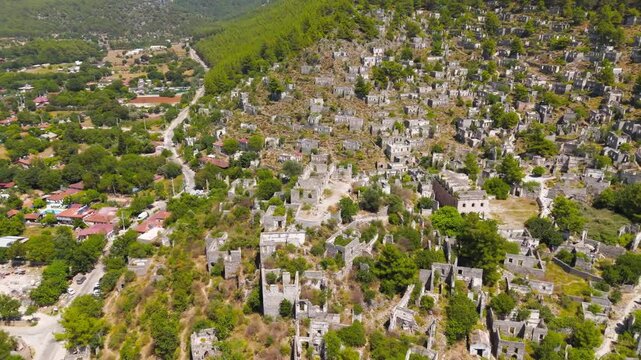 Kayakoy, Turkey. Aerial drone view of Turkish flag waving over abandoned ghost town ruins on rocky hillside slope under clear blue sky, with dense pine forest and distant mountain ridges. Aerial View