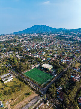 Aerial view of the Cihideung area featuring a green soccer field, surrounding residential houses, lush trees, and a prominent mountain in the background in Parongpong, West Java, Indonesia.