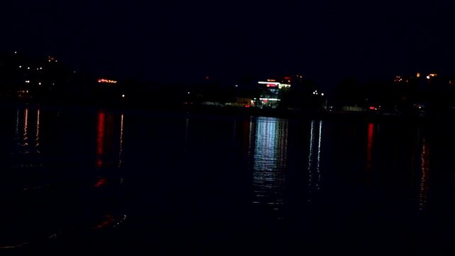 Shimmering lights cast colorful patterns over calm waters. The evening skyline rests beneath dark skies. Waterfront views shine in Udaipur, Rajasthan.