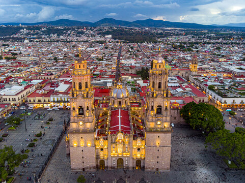 Aerial view of Morelia Cathedral illuminated at dusk, surrounded by the historic city center, Plaza de Armas, colonial architecture, and distant mountains in Morelia, Michoacan, Mexico.