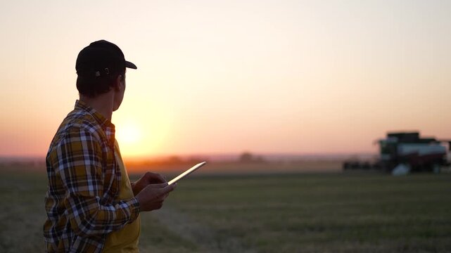 Farmer uses tablet checking field data at sunset. Agricultural worker reviews digital information on tablet. Farmer monitors crops using technology. Harvester works in field during golden hour.