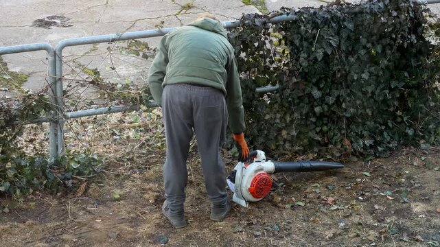 A gardener cleaning around a fence with a leaf blower, performing outdoor maintenance tasks to prepare a yard for the season while ensuring a tidy landscape in a residential area
