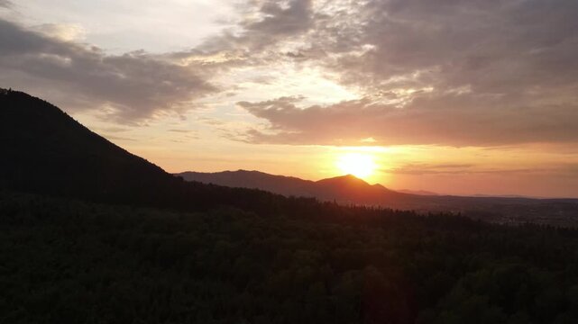 Aerial drone view of beautiful mountains at warm sunset. Forest slopes of mountain range. Bright disk of sun slowly sets tops of mountains. Dramatic sunrise sky background