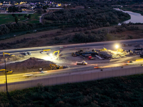 Aerial view of the 2100 North Freeway construction site at night, featuring heavy machinery, bright work lights, and traffic light trails near the Jordan River in Lehi, Utah, United States.