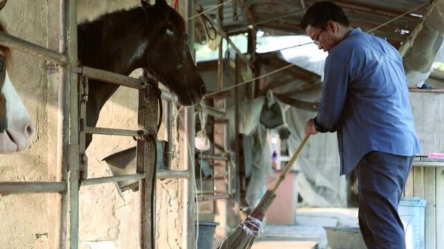 Male farmer sweeping dry straw in front horse stables where piles straw scattered all over ground : Farmer wielding broom works diligently attentively dedication keeping horse farm spotlessly clean.