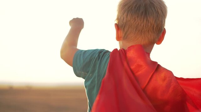 Boy wearing superhero cape playing outdoors at sunset. Child pretends to fly with arms raised. Kid in red cape plays hero. Young boy imagines flying. Childhood play and imagination at golden hour.