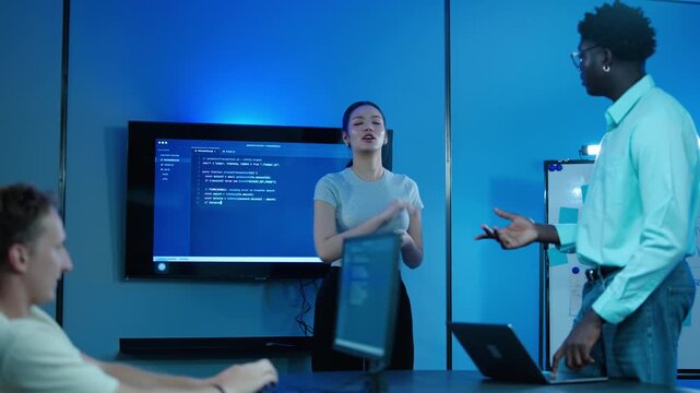 Diverse software engineering team collaborating on complex coding project in modern blue-lit tech office. Asian female lead programmer presents code on large monitor during a late night IT meeting.