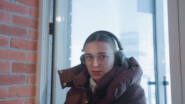 White woman at cafe door warming hands, heavy winter jacket and headphones, frosted glass and snow outside, brief pause before ordering, soft brick interior and muted morning light