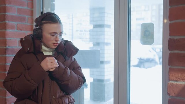 Caucasian woman warms hands inside cafe, commuter pressing palms together for heat with headphones on, steam from nearby cup, brick wall backdrop and frosty window view, quiet relieved smile