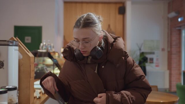 Young person at cafe counter adjusting headphones and browsing display, checking phone then ordering pastry and drink, warm pendant lights and plants, walking through seating area to meet friend