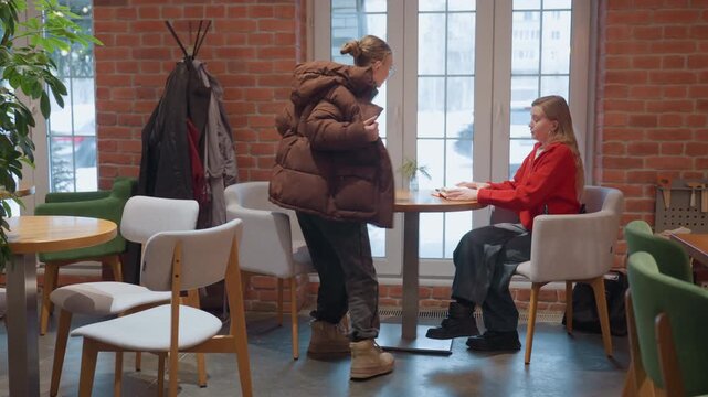 White women share cafe conversation and coffee. Two friends sit across round table near window, laugh and gesture, cozy brick interior with plants and coat rack, mugs and pastry on table, warm