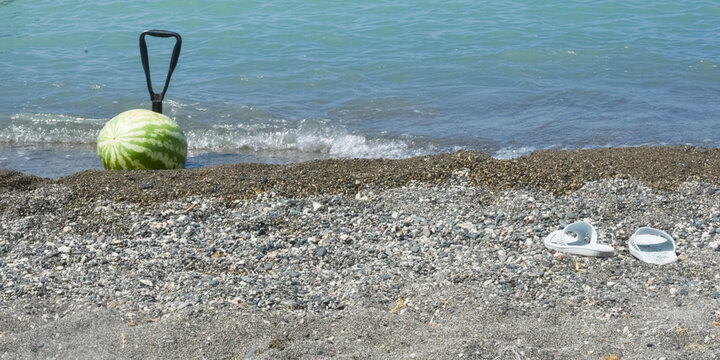 Whole green watermelon in net bag anchored in sea water with shovel handle on dark pebble beach shore with shoes in foreground and copy space