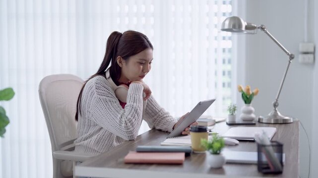 Young Asian woman using digital tablet for online education course at modern home workspace
