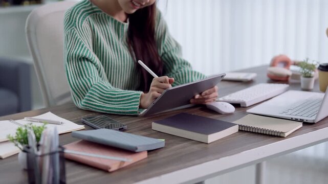 Young Asian woman writing notes on digital tablet with stylus at organized home workspace for education