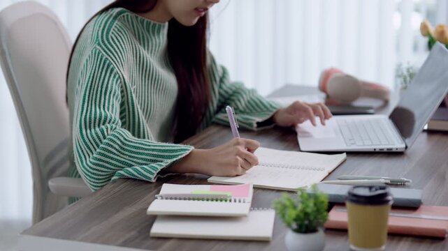 Young Asian student writing notes from laptop during online learning session at bright home desk