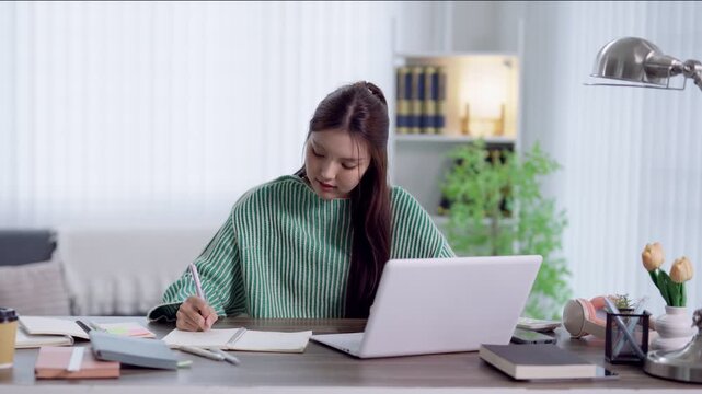 Young Asian student writing notes in notebook while studying with laptop at home desk