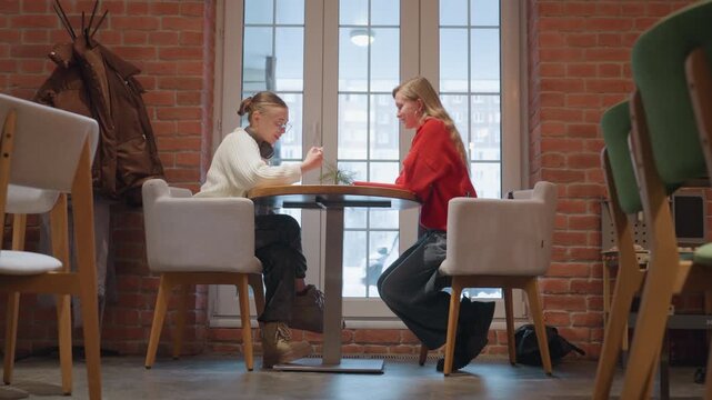 Two white women in cafe table reviewing notes and sketches, intimate mentorship between student and tutor with headphones, warm sunlight through window, exposed brick interior, soft conversation