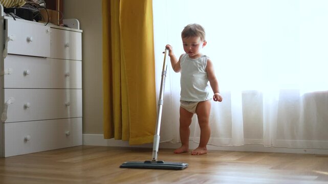 Adorable barefoot toddler in gray tank top and diaper holding a flat mop and cleaning wooden parquet floor at home. Bright room with yellow and white curtains and white dresser in background.