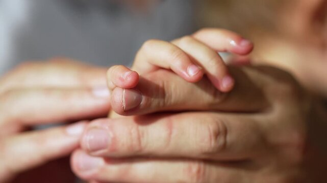 Adult parent hand gently holds tiny baby hand showing tender care. Large fingers carefully cradle small baby finger. Soft human skin shows deep family bond. Loving parent touch comforts infant.