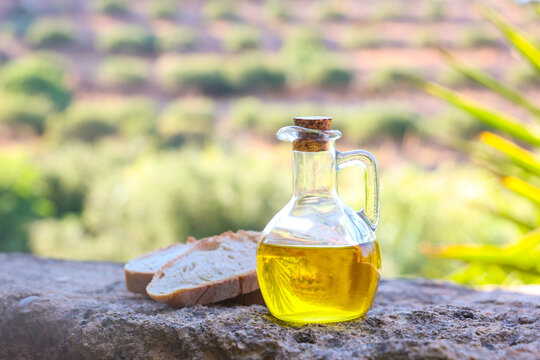 A rustic glass bottle of golden olive oil beside fresh bread on a stone ledge overlooking a sunlit olive grove in Crete, Greece, capturing Mediterranean tradition and natural rural beauty.
