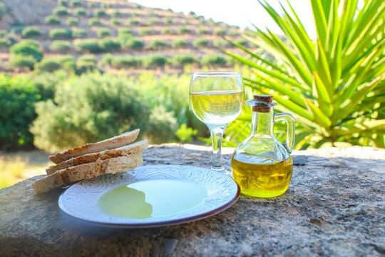 Olive oil, bread, and wine sit on a stone table overlooking terraced olive groves in Crete, Greece, celebrating Mediterranean cuisine and the harmony of food and landscape.