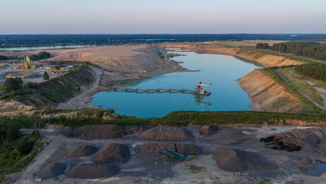 Aerial view of a large sand mine with a floating and fully automated dredger in a turquoise water basin, surrounded by sand piles and forest in Poland.