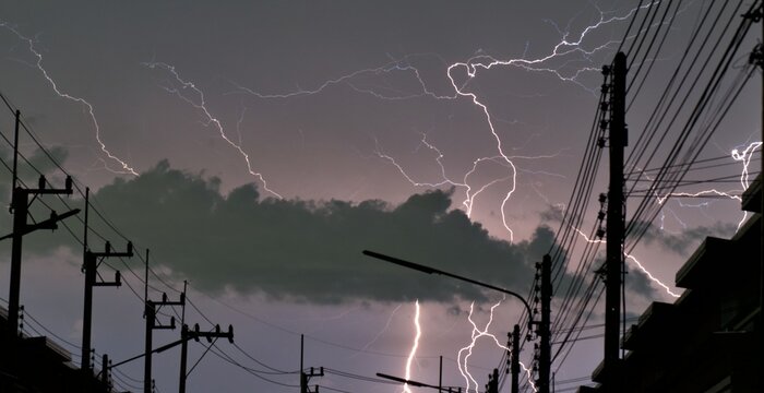 Widespread lightning storm over an urban street with power lines and utility poles silhouetted against a dramatic cloudy night sky.