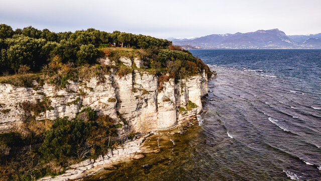 Aerial view of the rocky cliffs of Grotte di Catullo overlooking the blue waters of Lake Garda with lush trees and distant mountains in Sirmione, Lombardy, Italy.