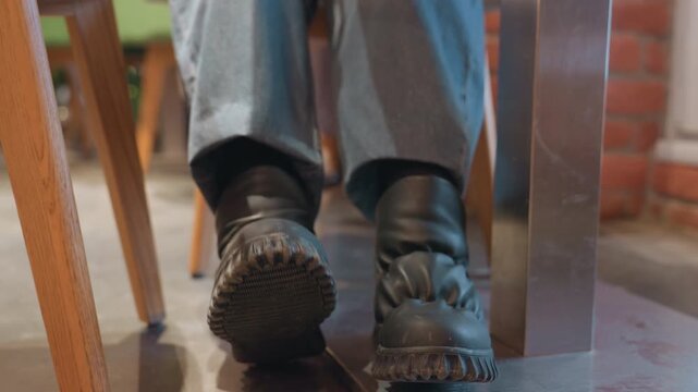 Black boots tapping under cafe table, impatient patron shuffling feet, businessperson checking watch, busy brick cafe ambiance, rhythmic sole tread, afternoon chatter and clatter, sense of hurry