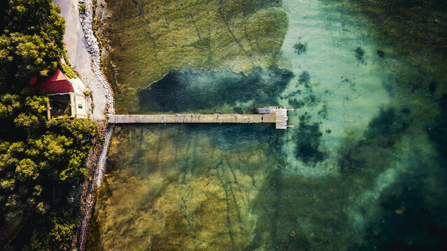 Aerial view of a wooden pier extending into the clear turquoise waters of Lake Garda, with a small building and lush trees along the shoreline in Sirmione, Lombardy, Italy.