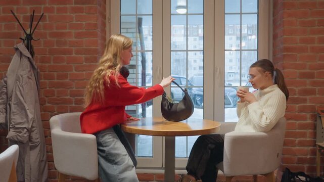 Intimate cafe table two white women share coffee and conversation, friend places purse and handbag on table then sits, gentle smiles and nods, casual winter coats by coat rack, warm ambience