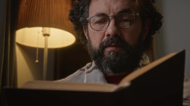 Middle aged Caucasian man with glasses reading book at home, turning pages. Video ideal for commercial projects focusing on mental wellbeing, digital detox, education, or leisure activities