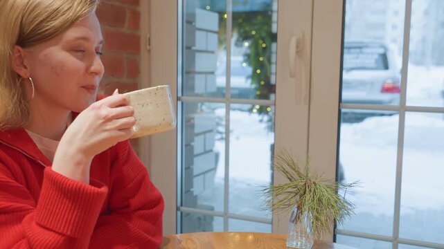 Caucasian woman with friend in cafe exchanging mugs near window, snow visible on street, sharing warm latte, red sweater, hand gesture toast, candid smile, cozy winter mood, urban bistro backdrop,