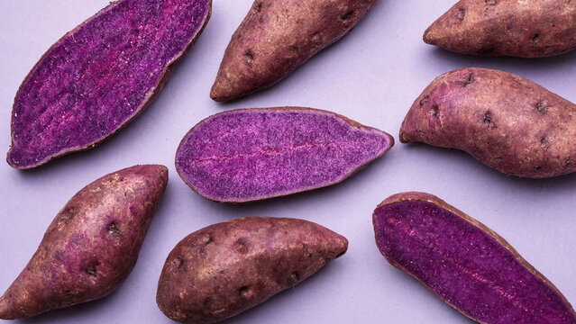 Flat lay composition of whole and longitudinally sliced purple sweet potatoes ube scattered across a clean lilac background