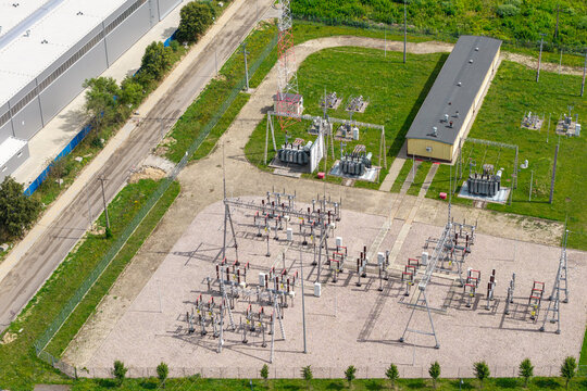 Aerial view of a small electrical substation featuring transformers, metal switchgear structures, and a transmission tower set on a gravel lot in Poland.