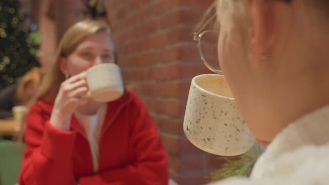 Two white friends toasting coffee at cafe table, red jacket and cream sweater contrast, ceramic mugs clink, relaxed conversation, soft brick background, candid laughter and warmth