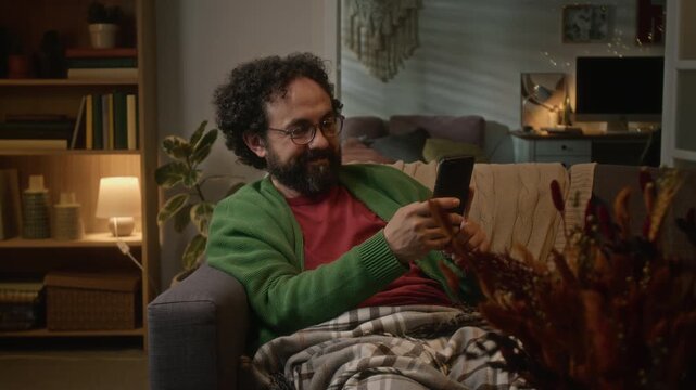 Middle aged Caucasian man sitting on couch, scrolling on smartphone and rotating it horizontally to watch video. Footage is perfect for mobile app promotions or streaming service advertisements