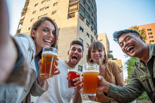 Joyful friends cheering with beer during a low angle city selfie. Diverse millennials and gen z adults laugh close to the camera outdoors. Happy hour concept